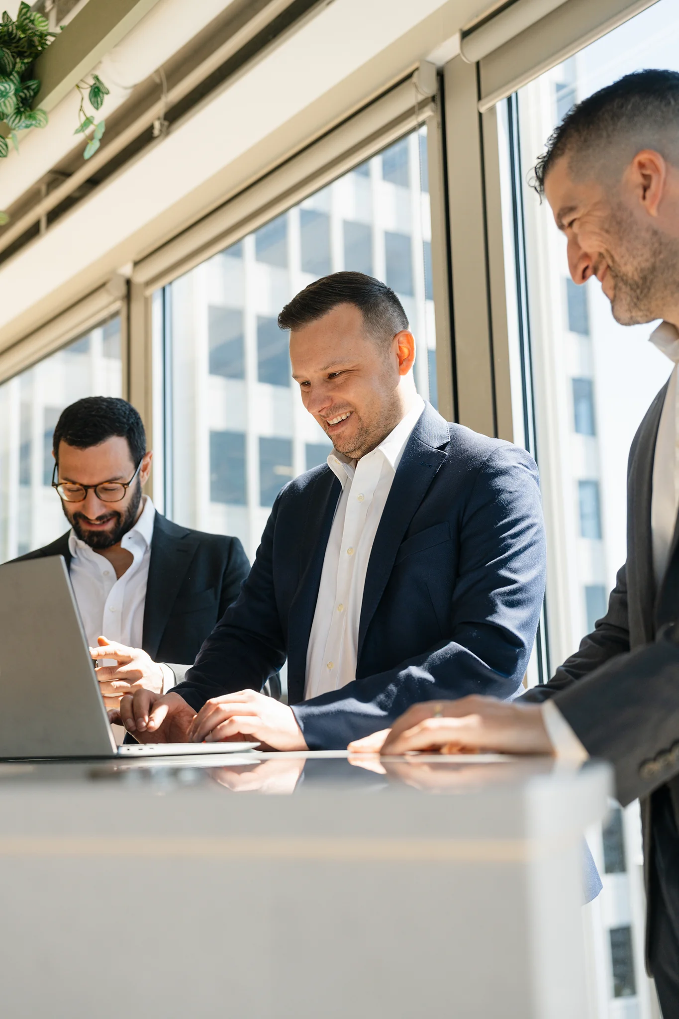 Trois professionnels en réunion debout autour d'un portable, lumière naturelle de bureau en hauteur.